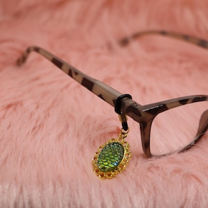 Puede incluir: Accesorio para gafas con un colgante dorado y ornamentado. El colgante tiene un diseño verde iridiscente en forma de escamas. El colgante está sujeto a unas gafas con montura marrón y beige.