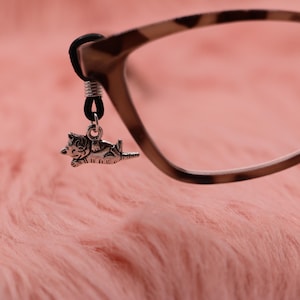 May include: A silver cat charm hangs from a pair of eyeglasses with a tortoiseshell pattern. The charm is attached to the glasses with a black rubber loop. The background is a soft, pink, fuzzy surface.