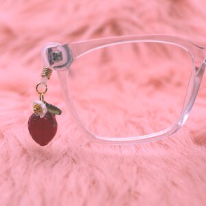 May include: Clear eyeglasses with a decorative strawberry charm. The charm is red with a white flower and green leaf accents, attached to the glasses frame with a gold-colored wire. The background is a soft, pink, textured surface.