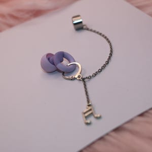 Op de afbeelding: Een unieke oorbel met een lavendel- en paars ontwerp. De oorbel heeft een ketting die verbinding maakt met een zilveren oormanchet. Een muzieknootbedel hangt aan de ketting. De oorbel ligt op een wit oppervlak.
