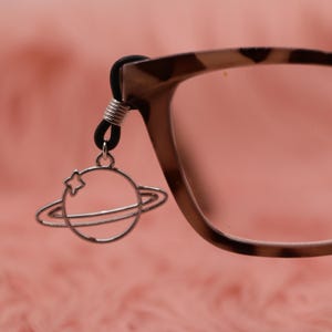 Puede incluir: Un dije plateado con forma de planeta y un acento de estrella cuelga de un soporte negro para gafas. El dije está sujeto a un par de gafas con un patrón de carey marrón y beige. El fondo es de color rosa suave.