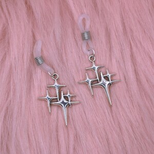 May include: Two silver eyeglass chains with starburst charms. The chains have clear rubber loops for attaching to glasses and silver coiled accents. The starburst charms have multiple points and are suspended from small silver rings. The background is a soft pink.