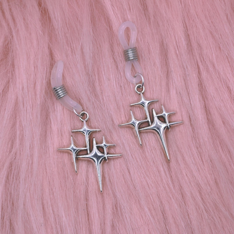 May include: Two silver eyeglass chains with starburst charms. The chains have clear rubber loops for attaching to glasses and silver coiled accents. The starburst charms have multiple points and are suspended from small silver rings. The background is a soft pink.