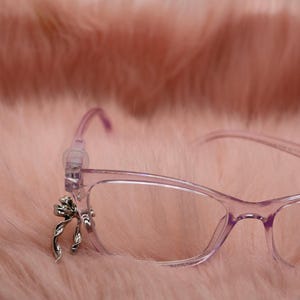 Może przedstawiać: Para jasnofioletowych okularów z srebrną kokardą na oprawce. Okulary są umieszczone na miękkim, różowym, futrzanym tle, tworząc delikatną i kobiecą estetykę.