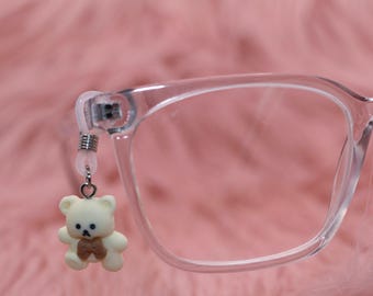 May include: Clear eyeglasses with a small, dangling teddy bear charm. The bear is off-white with a brown bow tie. The glasses are set against a soft, pink background, creating a cute and playful aesthetic. The charm is attached to the glasses' arm.