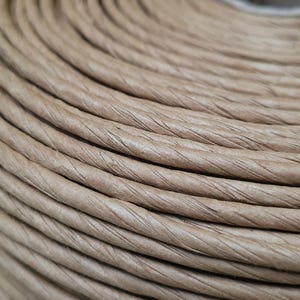 Pode incluir: Close-up de um rolo de barbante de papel de cor natural. O barbante é enrolado firmemente, criando círculos concêntricos. A textura do barbante é visível, com variações sutis de cor e uma superfície ligeiramente áspera.