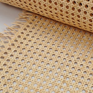 May include: A roll of light tan natural cane webbing featuring a classic octagonal woven pattern. The material displays a consistent grid of small holes formed by interlaced strips of plant fiber, with frayed edges visible at the bottom.