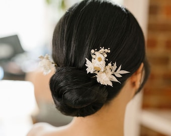 May include: A woman's dark hair is styled in a low bun, adorned with delicate white floral hair accessories. The arrangement includes small, clustered blooms and larger, petal-like elements, creating an elegant, natural look for weddings or special events.