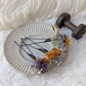 Puede incluir: Tres horquillas negras adornadas con flores secas en tonos naranja, morado y blanco. Los accesorios florales para el cabello están dispuestos sobre un plato crema con diseño acanalado, sobre un fondo blanco y esponjoso.