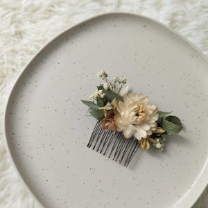 Puede incluir: Un peine floral para el cabello con una flor grande de color crema y flores más pequeñas rosas y blancas. Hojas verdes y delicados acentos blancos están dispuestos en un peine negro, perfecto para bodas u ocasiones especiales.