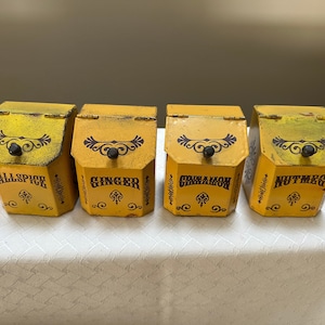 May include: Four yellow metal spice jars with black lids and brown lettering. The jars are labeled "Allspice", "Ginger", "Cinnamon", and "Nutmeg".