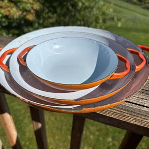 May include: A set of three round, nested enamel pans with orange exteriors and light blue interiors. Each pan features two curved orange handles on opposite sides. The edges of the pans show signs of wear with dark, distressed metal rims.