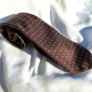 May include: A brown tie with a repeating pattern of small white and blue floral designs and paisley shapes. The tie is made of a textured fabric and is lying on a white surface.