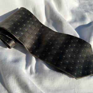 May include: A dark gray tie with a subtle checkered pattern. The tie is made of a textured fabric and is folded over itself. The tie is lying on a white, textured surface.