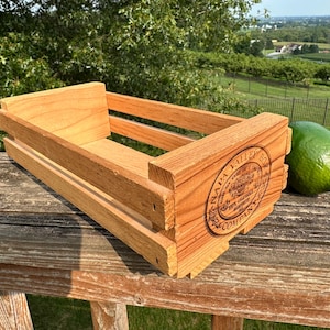 May include: A rectangular wooden crate with open slats and a natural light brown finish. The front panel features a circular black stamp that reads Napa Valley Box Company. A single green lime is positioned next to the crate on a wooden surface.
