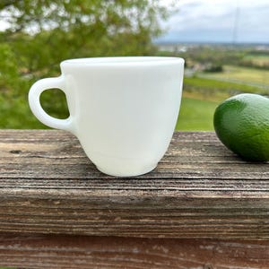 May include: A white ceramic teacup with a handle sits on a wooden surface next to a green lime. The teacup has a rounded shape and a smooth, glossy finish. The lime is a vibrant green color.