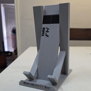 May include: A gray 3D-printed phone stand featuring the word "INTERSTELLAR" at the base and "TARS" on the main body. The stand has a unique, angular design.