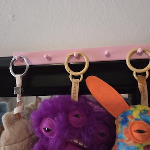 May include: A pink key rack with several plush toys hanging from it. The toys include a purple monster with a toothy grin, a tan bear, and a colorful creature with an orange ear. The keychains have the word "Fuggler" on them.
