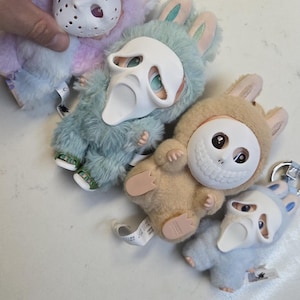 May include: A collection of plush bunny figures in various pastel colors, each wearing a white mask with different designs. The figures have soft, furry bodies and small feet. Some have keychains attached.