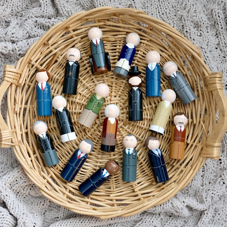 May include: A selection of wooden peg dolls, each painted to resemble figures in suits and coats, presented in a woven basket. The dolls display painted details, including hair and facial features, with suits in blue, black, and grey.
