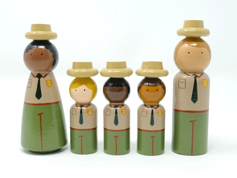 May include: Five wooden peg dolls depicting park rangers. Each doll wears a tan hat, a tan shirt with a green tie, and green trousers. The dolls vary in height and skin tone. The tallest doll is at the end of the line.
