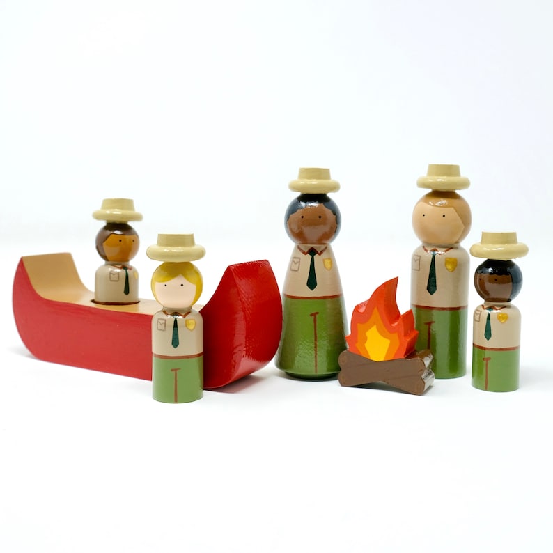 May include: A wooden toy set depicting a camping scene. It includes a red canoe with two figures, and several park ranger figures around a campfire. The figures wear tan hats, green skirts, and tan shirts. The campfire has orange flames.