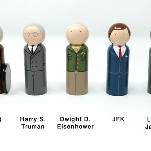 May include: Wooden peg dolls representing U.S. presidents: FDR, Harry S. Truman, Dwight D. Eisenhower, JFK, and Lyndon Johnson. Each doll is painted with details like suits, ties, and hair. FDR is in a wheelchair. Text labels identify each president.