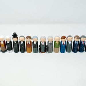 May include: A row of wooden peg dolls, each depicting a historical figure. The dolls are dressed in suits and other period attire, with painted details. The dolls are arranged in a line against a plain white backdrop.