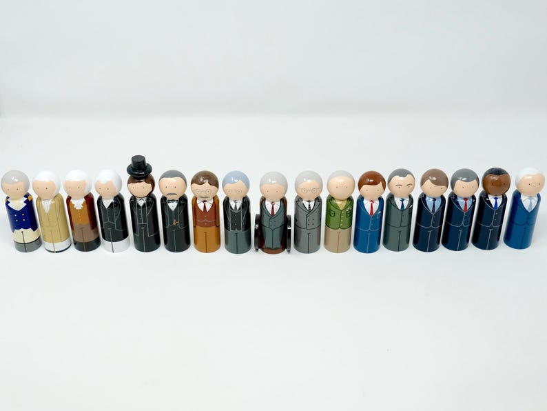 May include: A row of wooden peg dolls, each depicting a historical figure. The dolls are dressed in suits and other period attire, with painted details. The dolls are arranged in a line against a plain white backdrop.