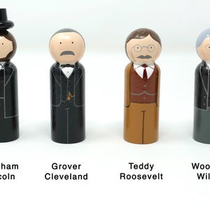 May include: Four wooden peg dolls representing U.S. presidents: Abraham Lincoln, Grover Cleveland, Teddy Roosevelt, and Woodrow Wilson. Each doll is painted with details of their attire, including suits, ties, and facial features. The names of the presidents are printed below.