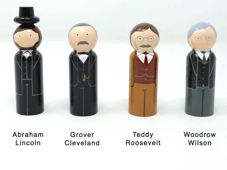 May include: Four wooden peg dolls representing U.S. presidents: Abraham Lincoln, Grover Cleveland, Teddy Roosevelt, and Woodrow Wilson. Each doll is painted with details of their attire, including suits, ties, and facial features. The names of the presidents are printed below.