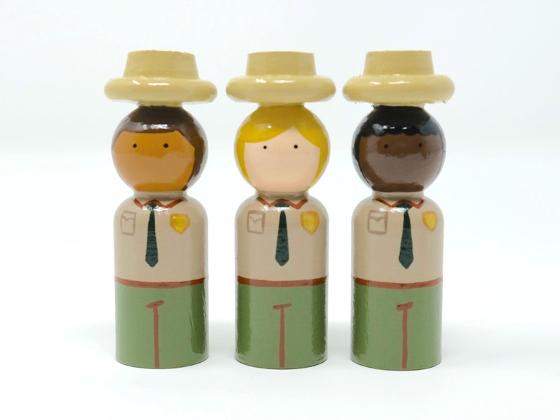 May include: Three wooden peg dolls depicting park rangers. Each doll has a different skin tone, blonde, brown, and dark brown. They wear tan shirts, green trousers, and tan hats. The dolls have painted details like ties and badges.