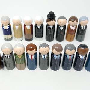 May include: A set of wooden peg dolls, each depicting a person in formal wear. The dolls display diverse skin tones, hairstyles, and suit colours, such as black, grey, blue, and brown. Some dolls have ties, and one is wearing a top hat.