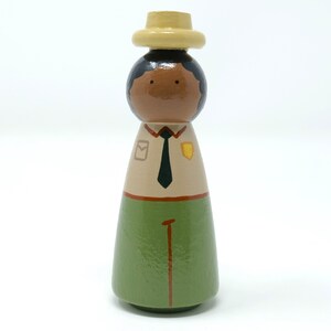 May include: A hand-painted wooden peg doll of a park ranger. The doll features a light brown face, black hair, and is dressed in a tan shirt, green trousers, and a pale yellow hat. The shirt has a black tie and badges.