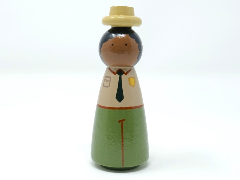 May include: A hand-painted wooden peg doll of a park ranger. The doll features a light brown face, black hair, and is dressed in a tan shirt, green trousers, and a pale yellow hat. The shirt has a black tie and badges.