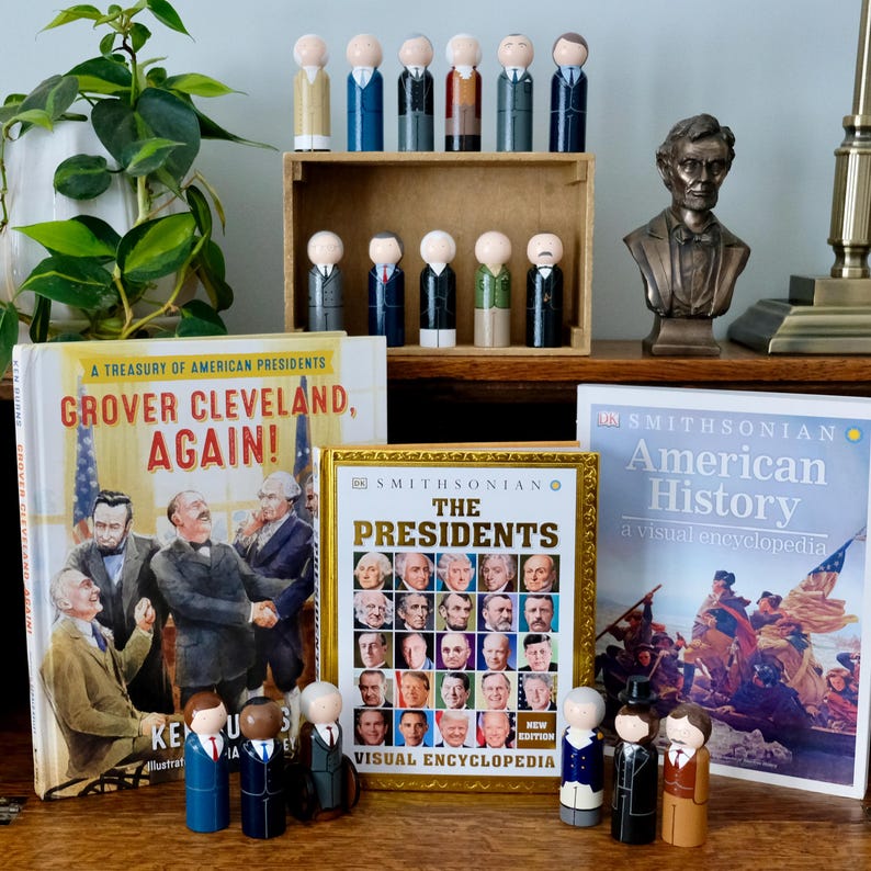 May include: A collection of wooden peg dolls depicting U.S. presidents, displayed with books on American history. The books include "Grover Cleveland, Again!" and "The Presidents: Visual Encyclopedia". The display includes a bust of Abraham Lincoln.