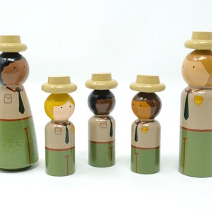 May include: A collection of wooden peg dolls representing park rangers. Each doll features a painted face, a beige hat, and a uniform with a green skirt or trousers. The dolls vary in height and skin tone, and are arranged together.