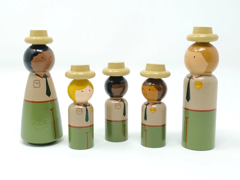 May include: A collection of wooden peg dolls representing park rangers. Each doll features a painted face, a beige hat, and a uniform with a green skirt or trousers. The dolls vary in height and skin tone, and are arranged together.