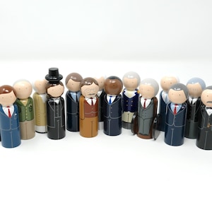 May include: A group of painted wooden peg dolls, each wearing a suit and tie. The dolls have different skin tones and suit styles, some with hats or briefcases. The figures are arranged together, possibly representing a meeting or a collection.