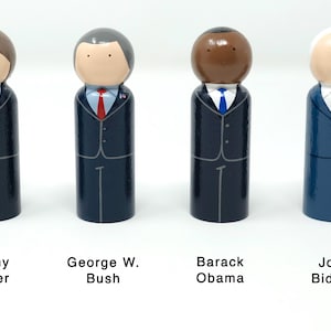 May include: Four wooden peg dolls representing U.S. presidents Jimmy Carter, George W. Bush, Barack Obama, and Joe Biden. Each doll is painted in a suit and tie, with the president's name below. The dolls have simple facial features.