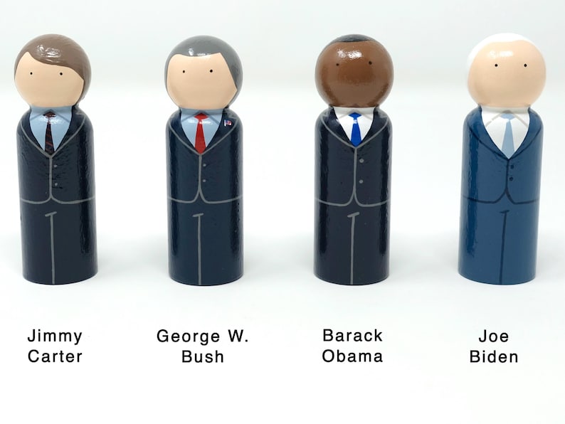 May include: Four wooden peg dolls representing U.S. presidents Jimmy Carter, George W. Bush, Barack Obama, and Joe Biden. Each doll is painted in a suit and tie, with the president's name below. The dolls have simple facial features.