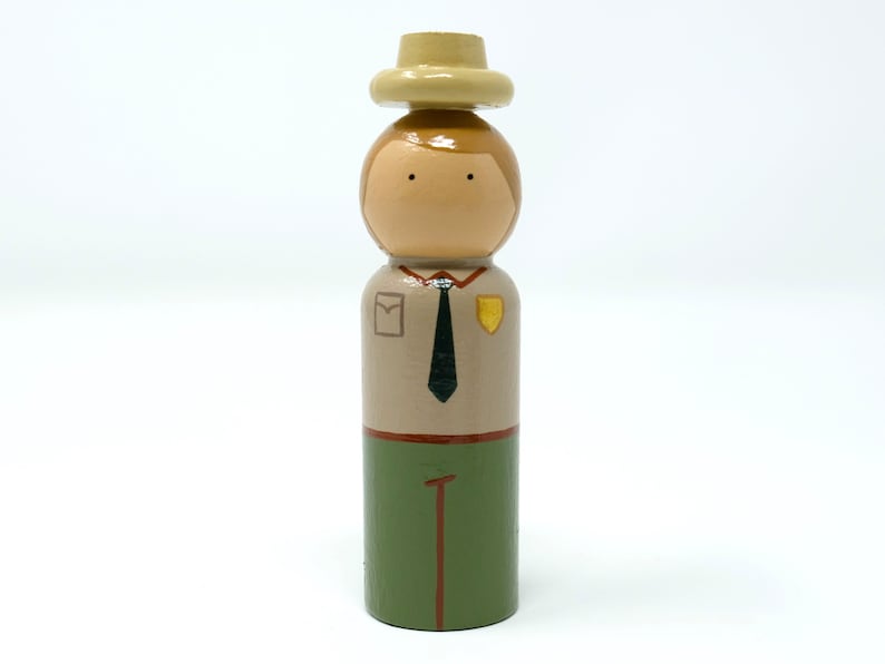 May include: Wooden peg doll of a park ranger. The doll features a tan hat, brown hair, and a tan uniform with a black tie, green trousers, and a badge. The doll is painted with simple details and stands upright.
