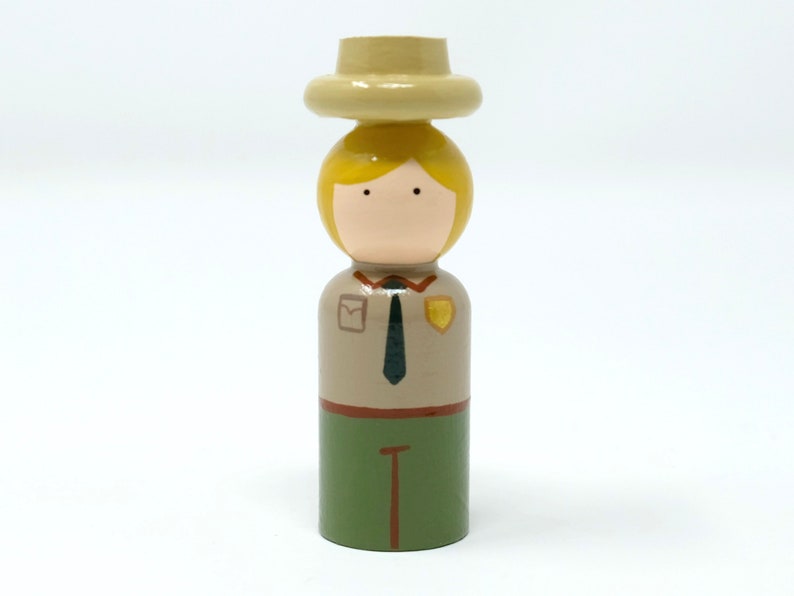 May include: A wooden peg doll of a park ranger. The doll features a tan hat, blonde hair, and a tan uniform with a green lower section. The doll has a black tie and a badge.
