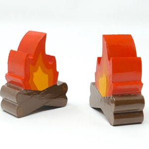May include: Two wooden campfire figurines. Each figurine features a stylized flame in shades of red and yellow atop two brown wooden logs. The figurines are painted with glossy paint and are set against a white background.