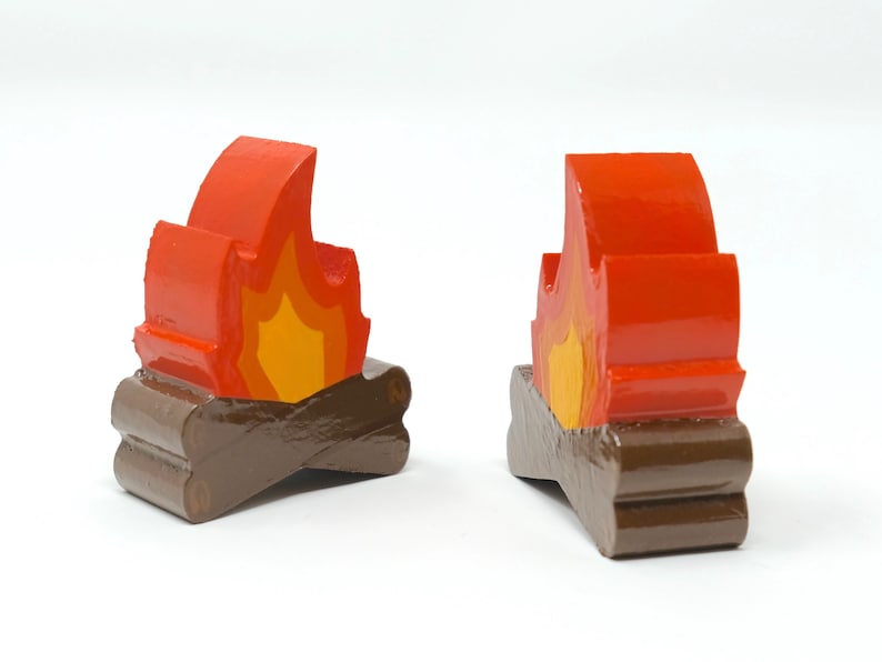May include: Two wooden campfire figurines. Each figurine features a stylized flame in shades of red and yellow atop two brown wooden logs. The figurines are painted with glossy paint and are set against a white background.