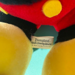 Puede incluir: Primer plano de un juguete de peluche Mickey Mouse amarillo con una etiqueta negra que dice "Disneyland Walt Disney World" y "Walt Disney Productions".