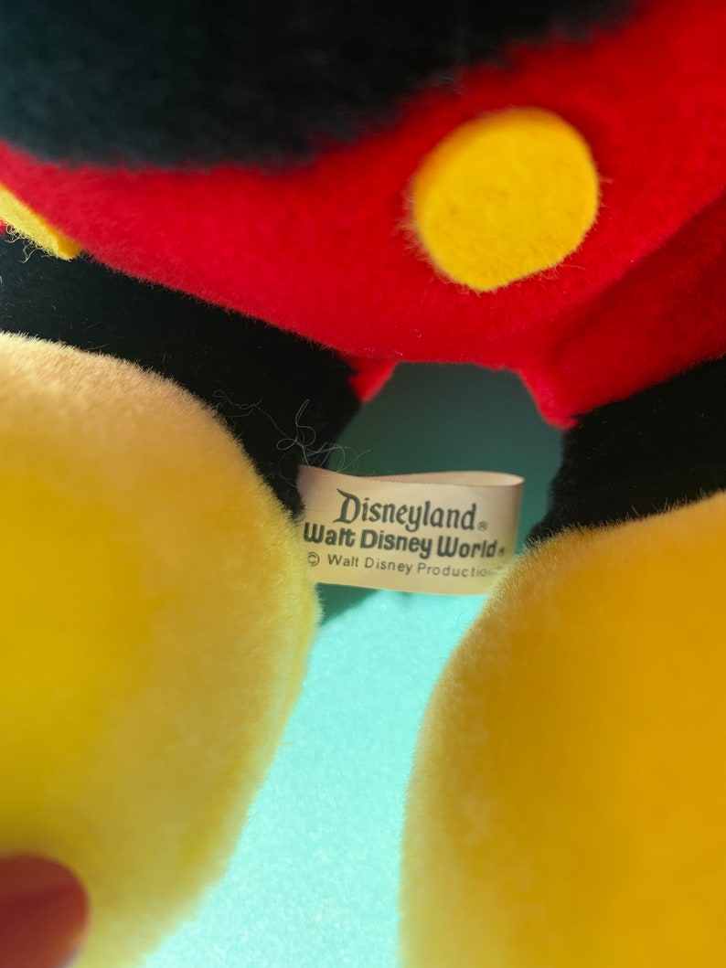 Puede incluir: Primer plano de un juguete de peluche Mickey Mouse amarillo con una etiqueta negra que dice "Disneyland Walt Disney World" y "Walt Disney Productions".