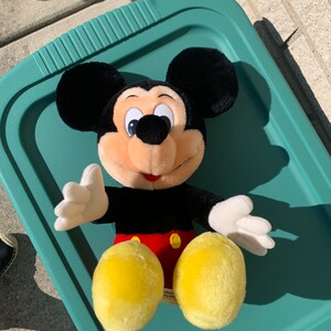Puede incluir: Un juguete de peluche Mickey Mouse negro y amarillo con un atuendo rojo y amarillo. El juguete est&aacute; sentado en una bandeja verde.