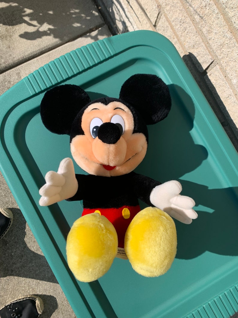Puede incluir: Un juguete de peluche Mickey Mouse negro y amarillo con un atuendo rojo y amarillo. El juguete est&aacute; sentado en una bandeja verde.