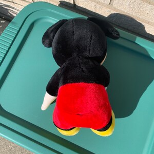 Puede incluir: Un juguete de peluche negro y rojo de Mickey Mouse. El juguete est&aacute; acostado boca abajo con la cabeza girada hacia un lado. El juguete lleva zapatos amarillos.